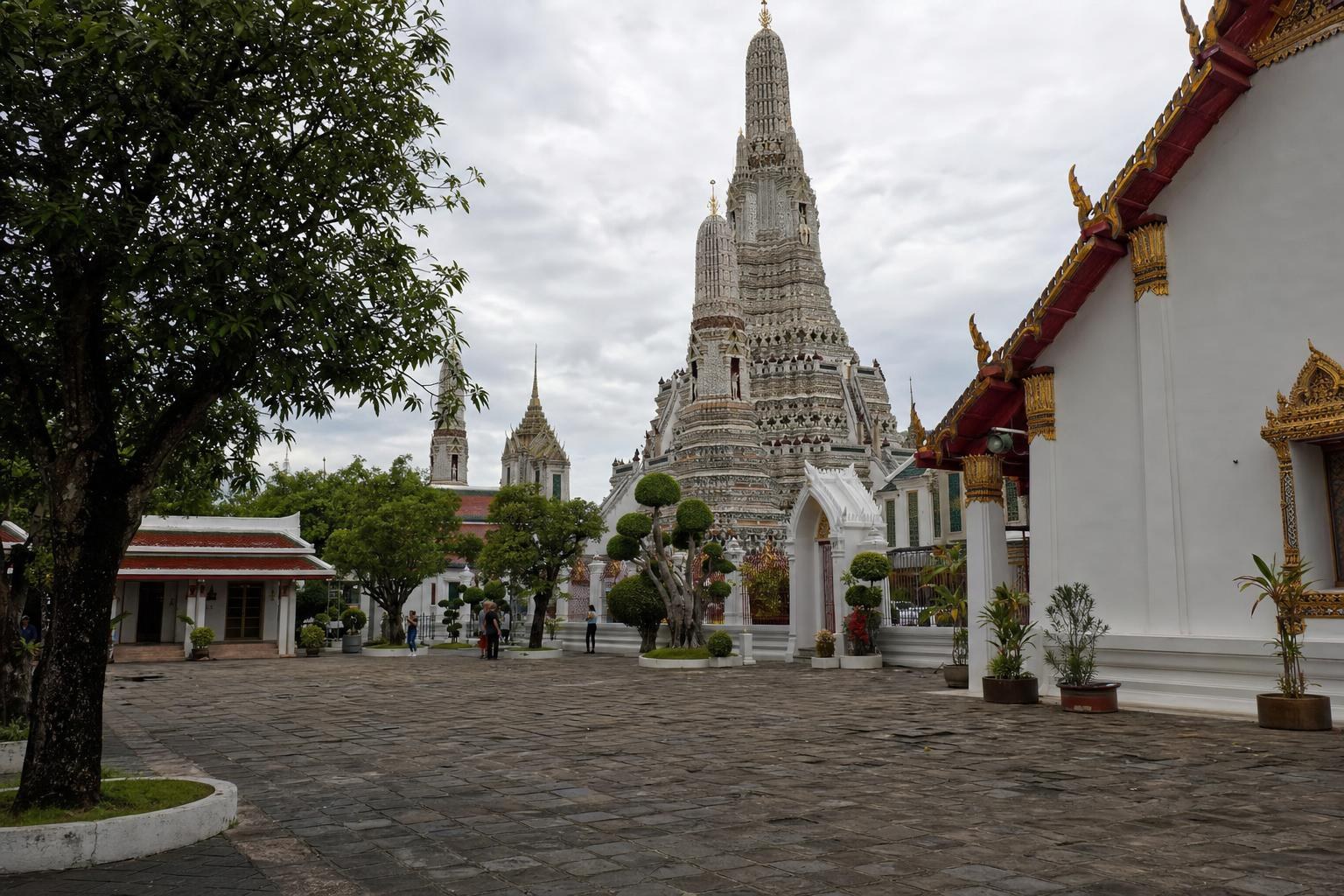 AI-generated illustration: Single ground-level view of Wat Arun temple courtyard in Bangkok, Thailand, ordinary natural daylight, authentic-looking weathered stone and white temple spire, one landmark with natural perspective, no skyline collage.