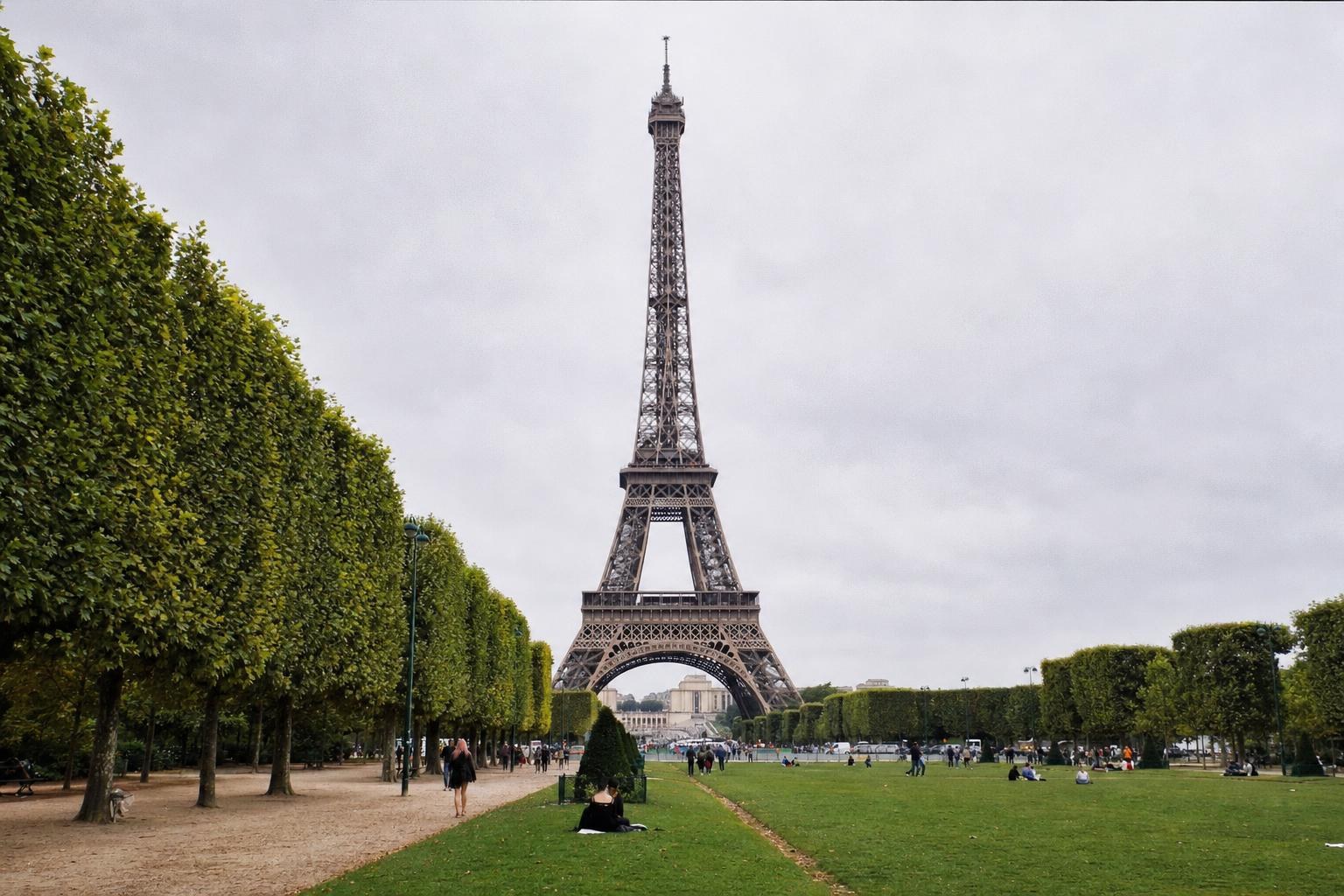 AI-generated illustration: The Eiffel Tower from the Champ de Mars park in Paris, France, eye-level viewpoint, ordinary grass and trees in foreground, overcast natural daylight, realistic iron structure and scale, no Louvre, Notre-Dame or other landmarks.
