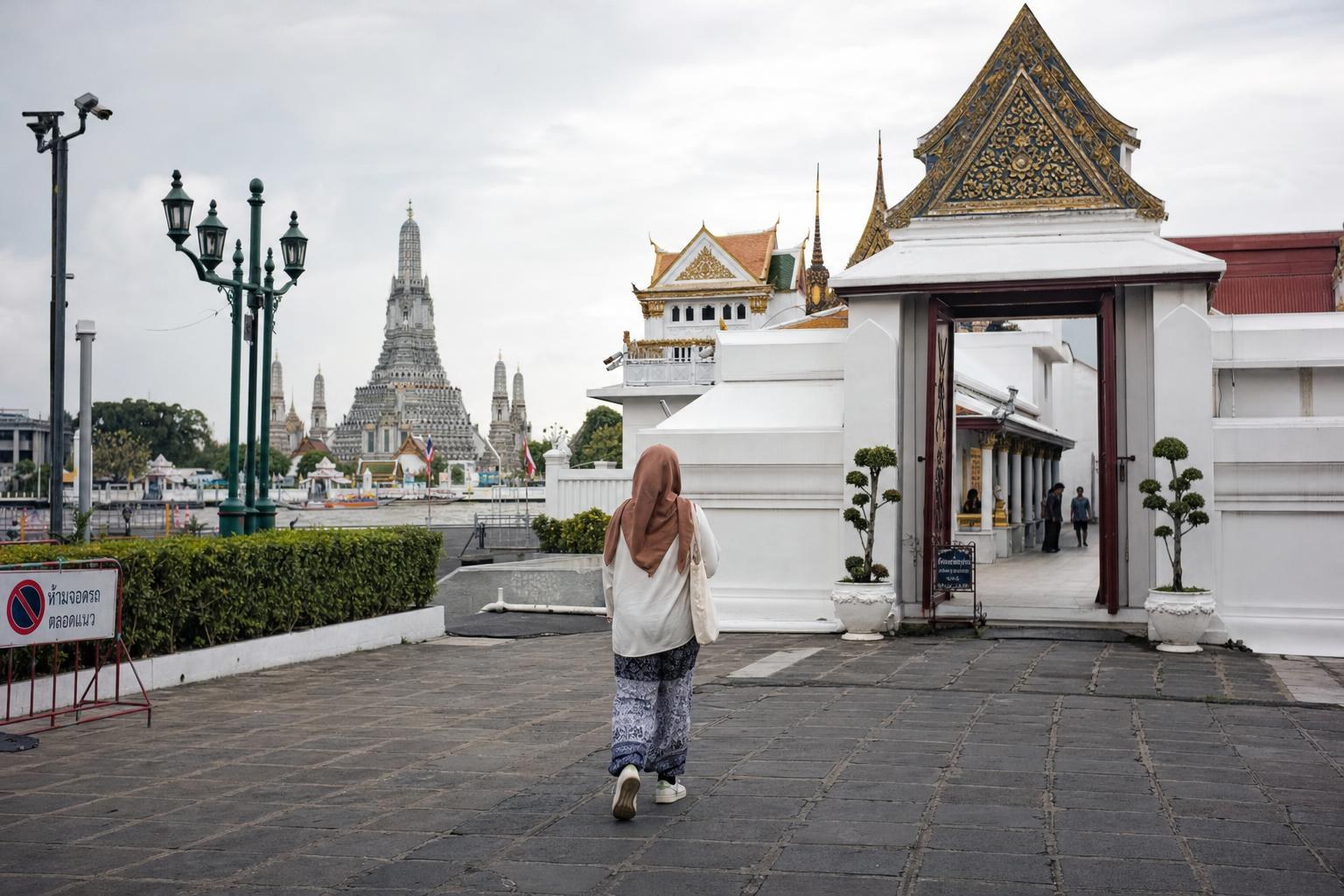 AI-generated illustration: A visitor in modest attire walking toward the Grand Palace entrance in Bangkok, Thailand, with river temple spires visible in the background